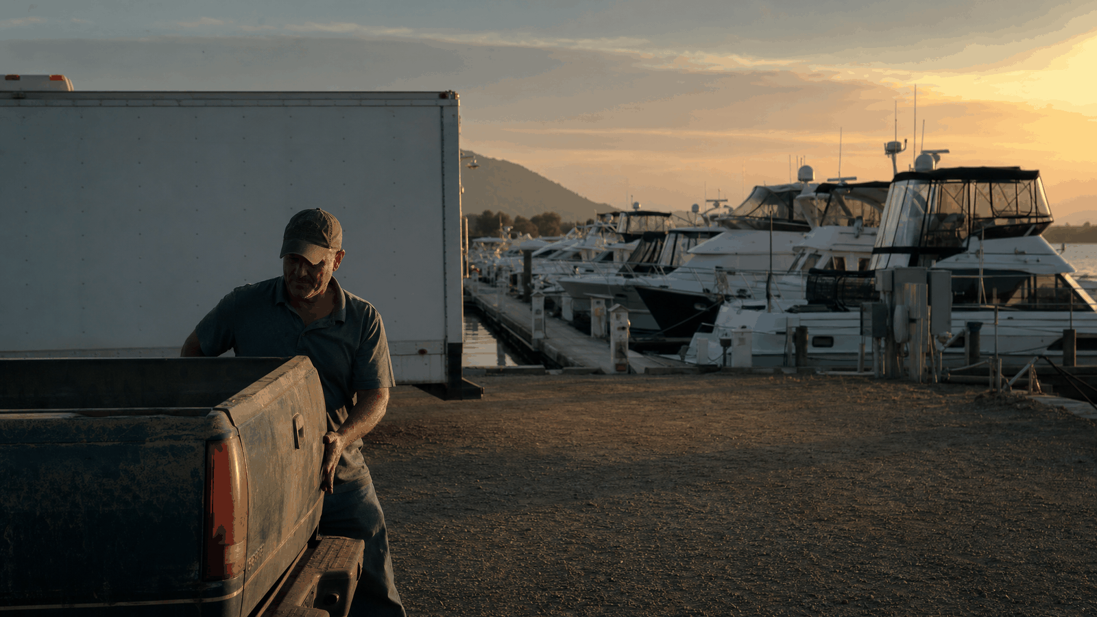 An inland lake marina at dusk, rows of large boats along floating docks, a middle-aged man in a polo shirt closing the tailgate of a pickup in the foreground